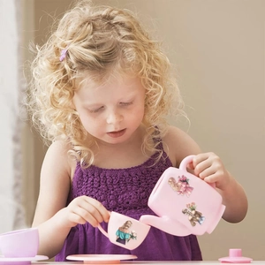 Toddler girl in purple dress playing with floral pink plastic tea set in a warm room