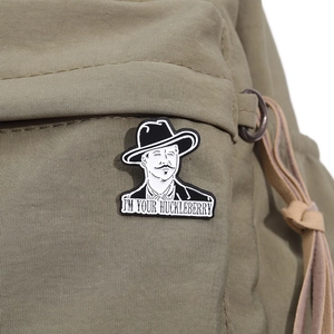 Close-up of cowboy enamel pin with I'M YOUR HUCKLEBERRY text on green canvas fabric