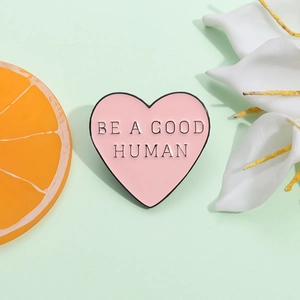 Spilla a cuore in smalto rosa minimalista con citazione Be A Good Human su sfondo menta