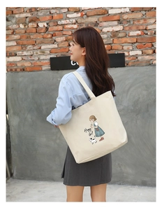 Cream canvas tote bag with Happy Girl cartoon print worn by a woman against a brick wall