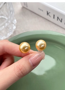 Detail of round golden pearl earrings with mirror-like luster held by a hand
