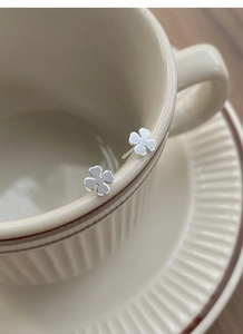 Minimalist silver four-leaf clover stud earrings resting on a vintage beige ceramic cup