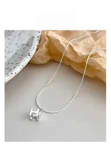 Gros plan du collier avec pendentif H en argent sur un fond beige minimaliste