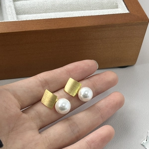 Detailed close-up of brushed gold square stud earrings with white pearls held by hand