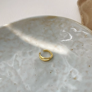 Minimalist gold X-shaped crisscross ear cuff on a textured ceramic dish with soft shadows