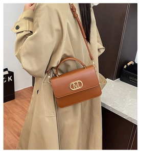 Detalle de bolso bandolera marrón vintage con hebilla M dorada sobre una gabardina beige