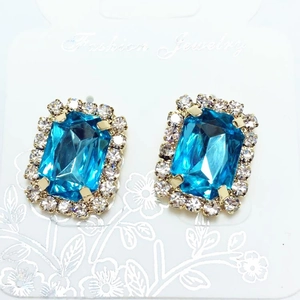 Close-up of light blue rectangular stud earrings with sparkling rhinestone halo and gold plating