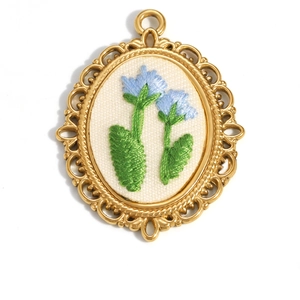 Detailed view of 18K gold floral oval pendant with blue embroidery on white fabric