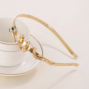 Elegant gold metal snake headband with green gemstone eyes on white ceramic background