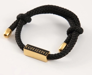 Detail eines schwarzen geflochtenen Armbands mit goldener Confidence Gravur auf Metall