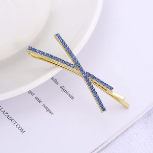 X-shape gold cross hair clip with deep blue rhinestones on a minimalist background
