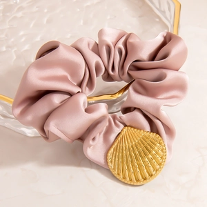 Roséfarbenes Satin-Haargummi mit goldenem Muschel-Anhänger auf einem Glastablett