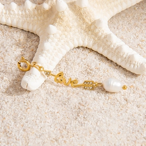 Detailed view of Love script gold pendant with baroque pearl and zircon on white sand