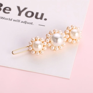 Eleganter goldener Haarclip mit drei weißen Perlenblüten auf einem sanft rosa Hintergrund