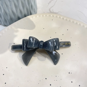 Close-up of a dark grey acetate hair clip featuring a layered 3D bow design and gold logo