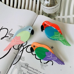Three colorful parrot-shaped cellulose acetate hair claws displayed on a magazine