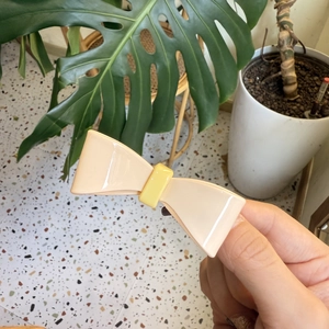 Cream-colored bow hair clip with yellow center accent shown in a hand-held lifestyle setting