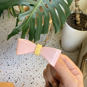 Pink bow hair clip with yellow center button shown in a natural minimalist setting