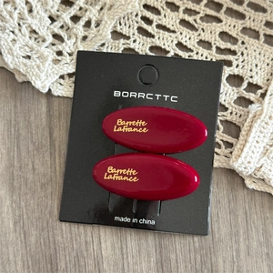 Detail showing two wine red acetate hair clips with gold logo on black display card