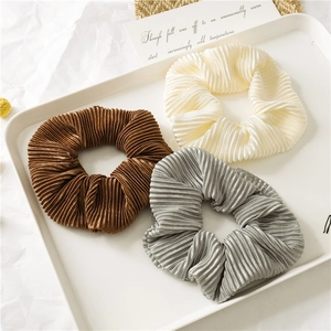 Close-up of three pleated satin scrunchies in brown, cream and silver on a white tray