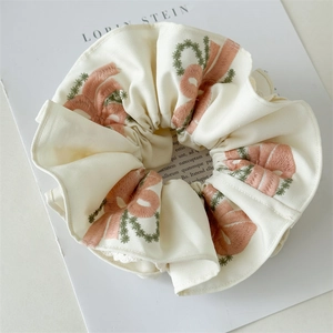 Close-up of cream white scrunchie featuring pink bow embroidery and green vine details