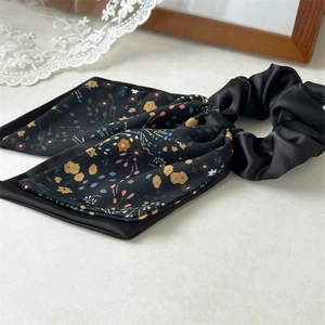Detail of a black satin scrunchie with a long floral ribbon on a light textured surface
