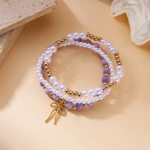 Set de tres pulseras con cuentas púrpuras, perlas blancas y dije de lazo dorado fino