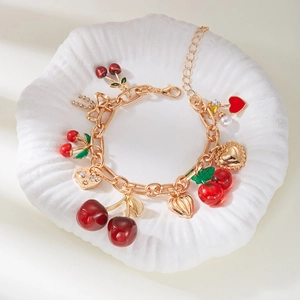 Close-up of golden paperclip necklace with red cherry, heart, and pearl charms on a shell