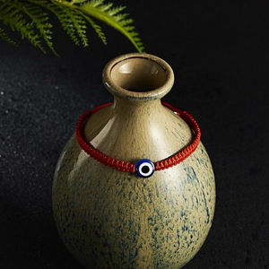 Close-up of a red woven Evil Eye bracelet displayed on a ceramic jar with green leaves