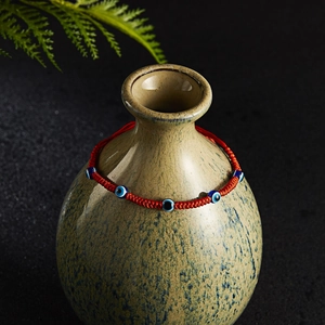 Macro detail of red woven Evil Eye bracelet on a vintage ceramic vase backdrop