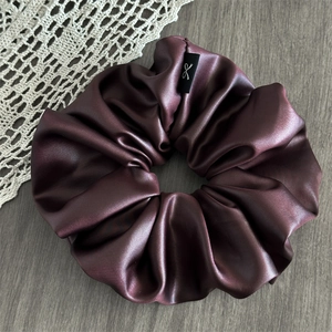 Macro detail of bordeaux satin scrunchie showing glossy texture and label on gray wood
