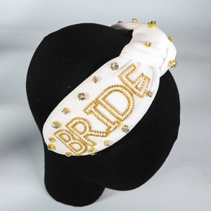 Detail eines weißen Knotenstirnbands mit goldener BRIDE Perlenstickerei und Strass