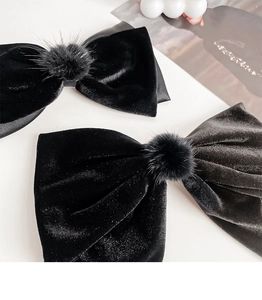 Detail of two black velvet bow hair clips with textured faux fur pom-poms on a white surface