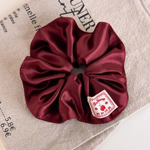 Close-up of wine red satin scrunchie with cherry brand label on beige canvas background