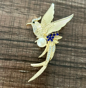 Macro detail of bird brooch showing golden feather textures, blue gems and a large white pearl