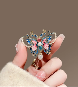 Main tenant une broche papillon en émail avec monture dorée, perles et pendentif perle