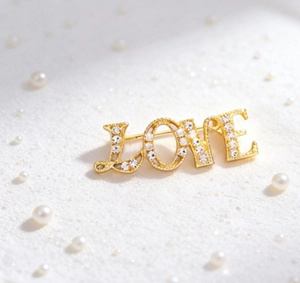 Close-up of golden LOVE brooch with inlaid crystals on white sand and pearls