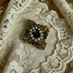 Dettaglio di una spilla vintage dorata con cristallo blu e perle su pizzo bianco