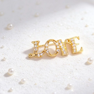 Elegant gold LOVE brooch with sparkling rhinestones and white pearls on a soft sand surface