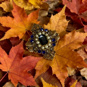 Macro di una spilla vintage dorata con pietra nera, perle e cristalli su foglie d'autunno