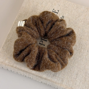 Close-up of deep brown plush scrunchie with brand tag on light textured linen surface