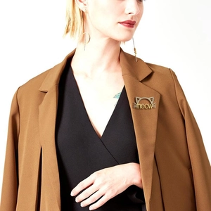 Détail d'une broche chat dorée avec texte meow sur un blazer marron avec boucles d'oreilles