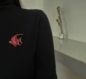 Detail einer roten Strass-Anstecknadel in Fischform auf einem schwarzen Rollkragenpullover