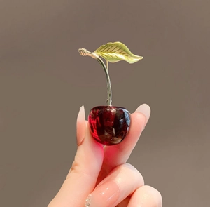 Detailed close-up of a red cherry brooch with golden leaves held between fingers