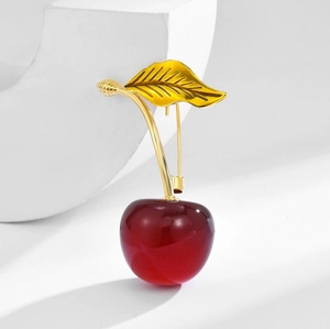 Elegant cherry brooch with red resin bead and golden leaf on a white geometric stand