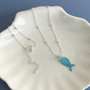 Detail view of a blue fish pendant on a silver beaded chain displayed on a shell dish