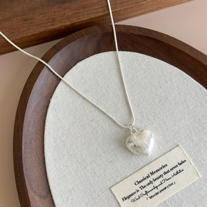 Detailed view of silver heart pendant with brushed texture on snake chain over linen