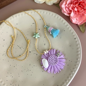 Détail du collier fleur violette tressée avec accents de coquillages et pompons bleus