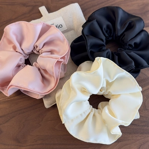 Detail of three pleated satin scrunchies in pink, black, and beige on a wooden surface