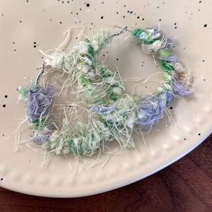 Dettaglio di orecchini a cerchio intrecciati con filato verde e viola su un piatto